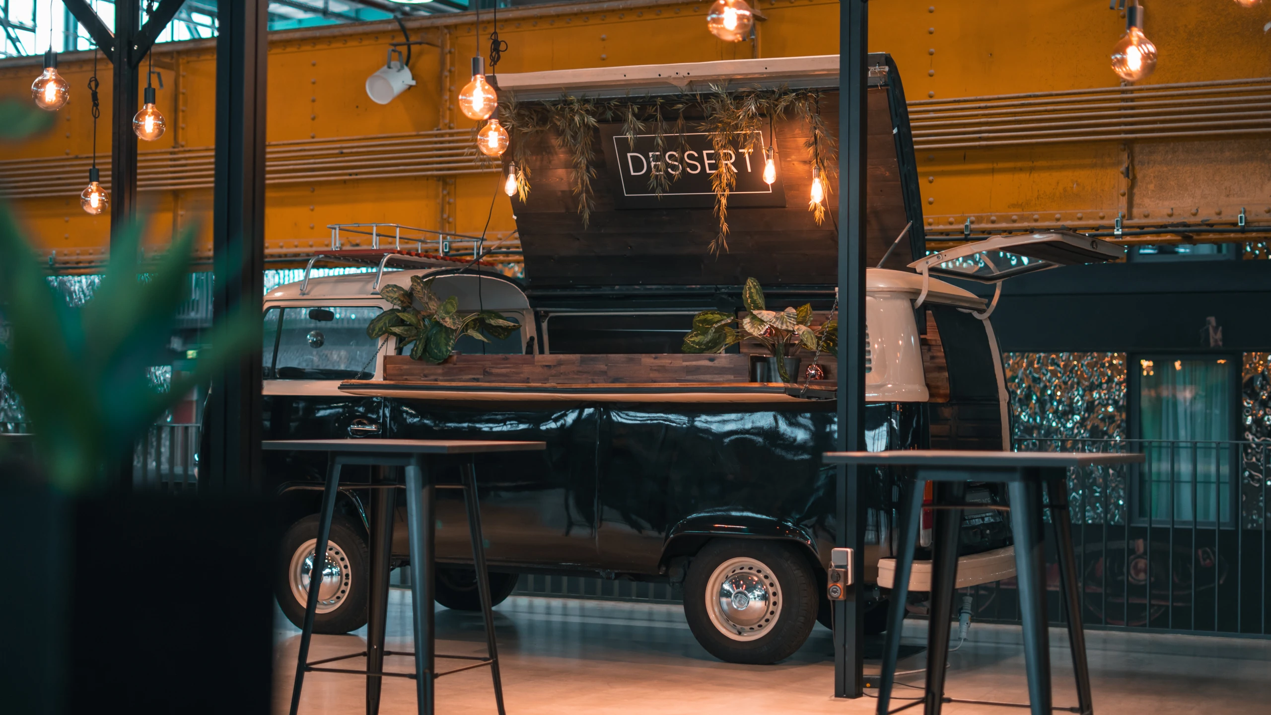 Ein stilvoller Dessertwagen, der an einen klassischen Transporter erinnert, steht im Mittelpunkt eines modernen Innenraums. Die Kombination aus gemütlichem Licht und Pflanzen schafft eine einladende Atmosphäre, ideal für süße Versuchungen. Vintage Dessertwagen in modernem Ambiente